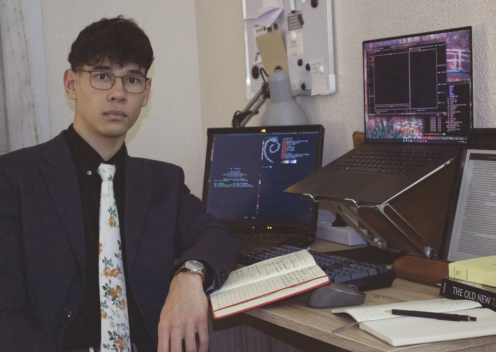 Black-haired mixed-race man in a suit in front of his desk, with computers.